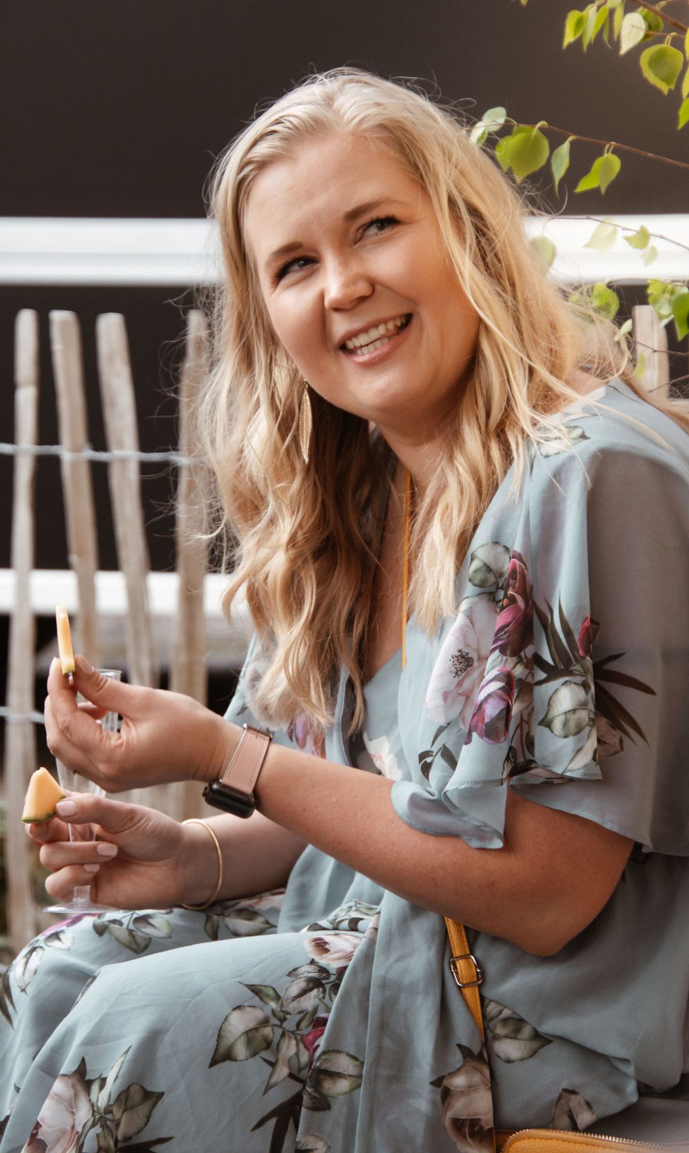 A woman in a floral dress sits outdoors smiling while holding a drink.