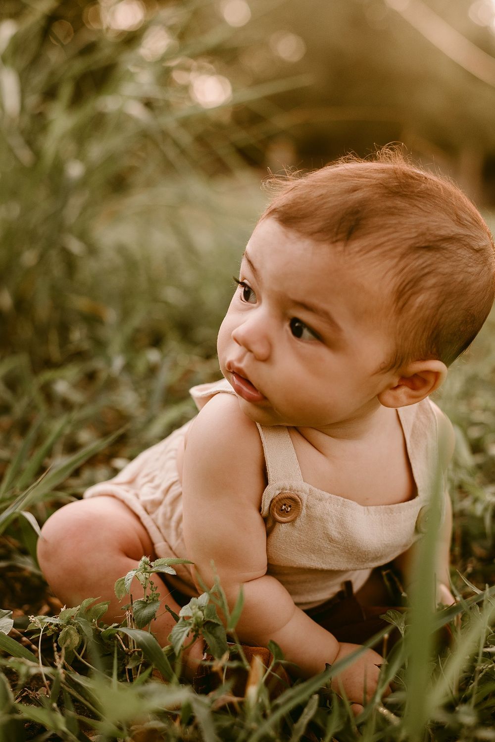 Baby in beige overalls looking up while sitting in green grass during sunset.