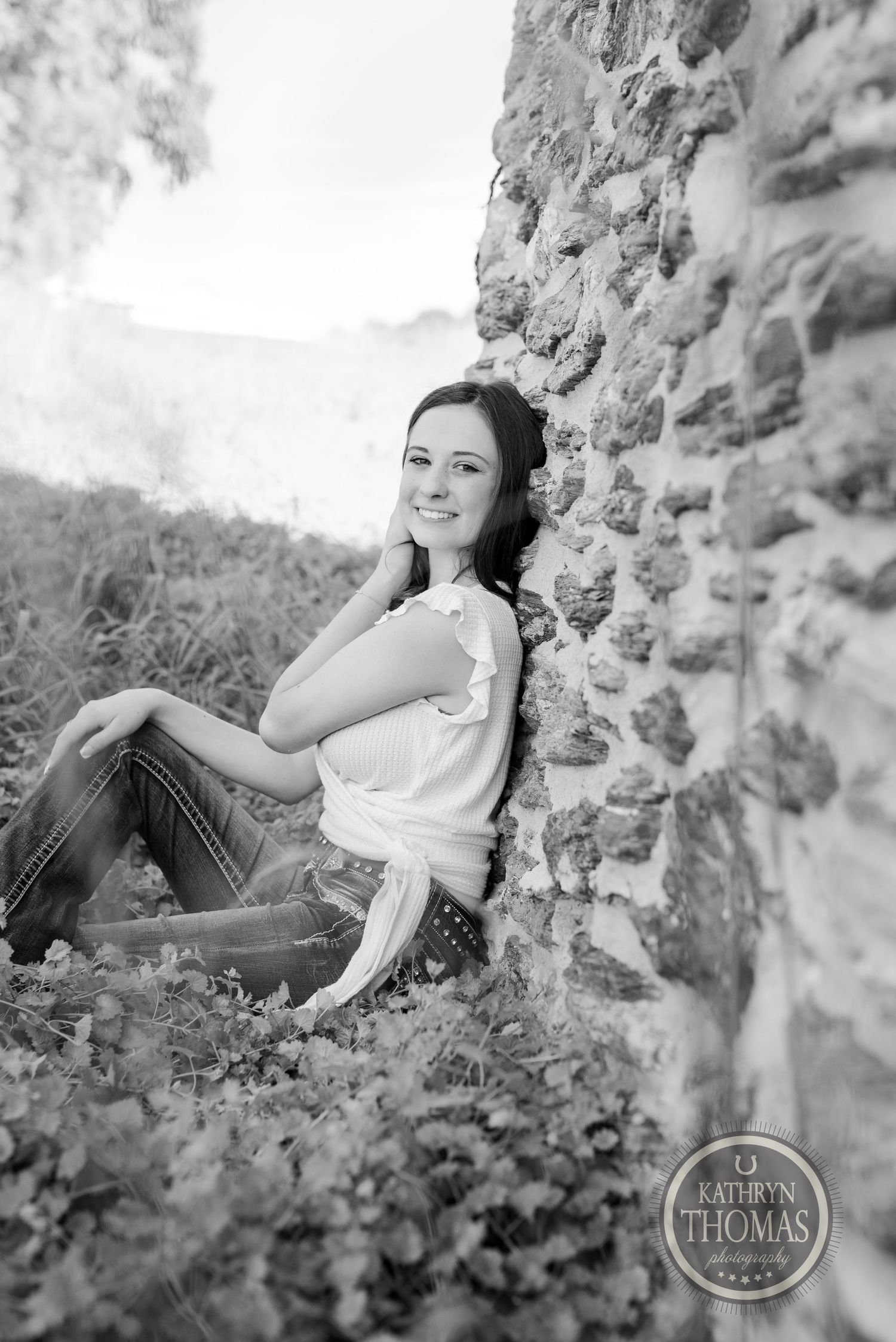 Black and white portrait of a woman sitting against a stone wall in nature.