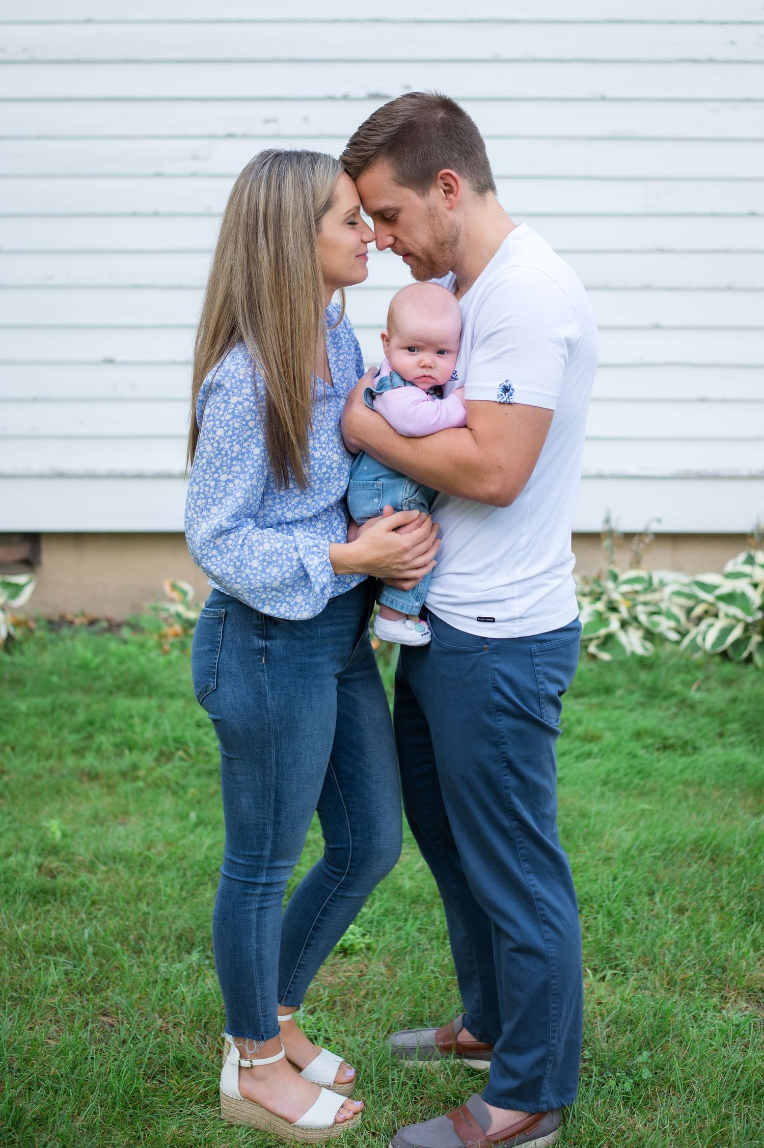 Young parents sharing an intimate moment while holding their baby against a white siding backdrop.