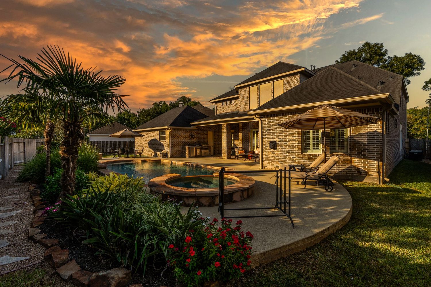 Luxury backyard patio with pool and stone features at sunset with dramatic orange sky and palm trees.