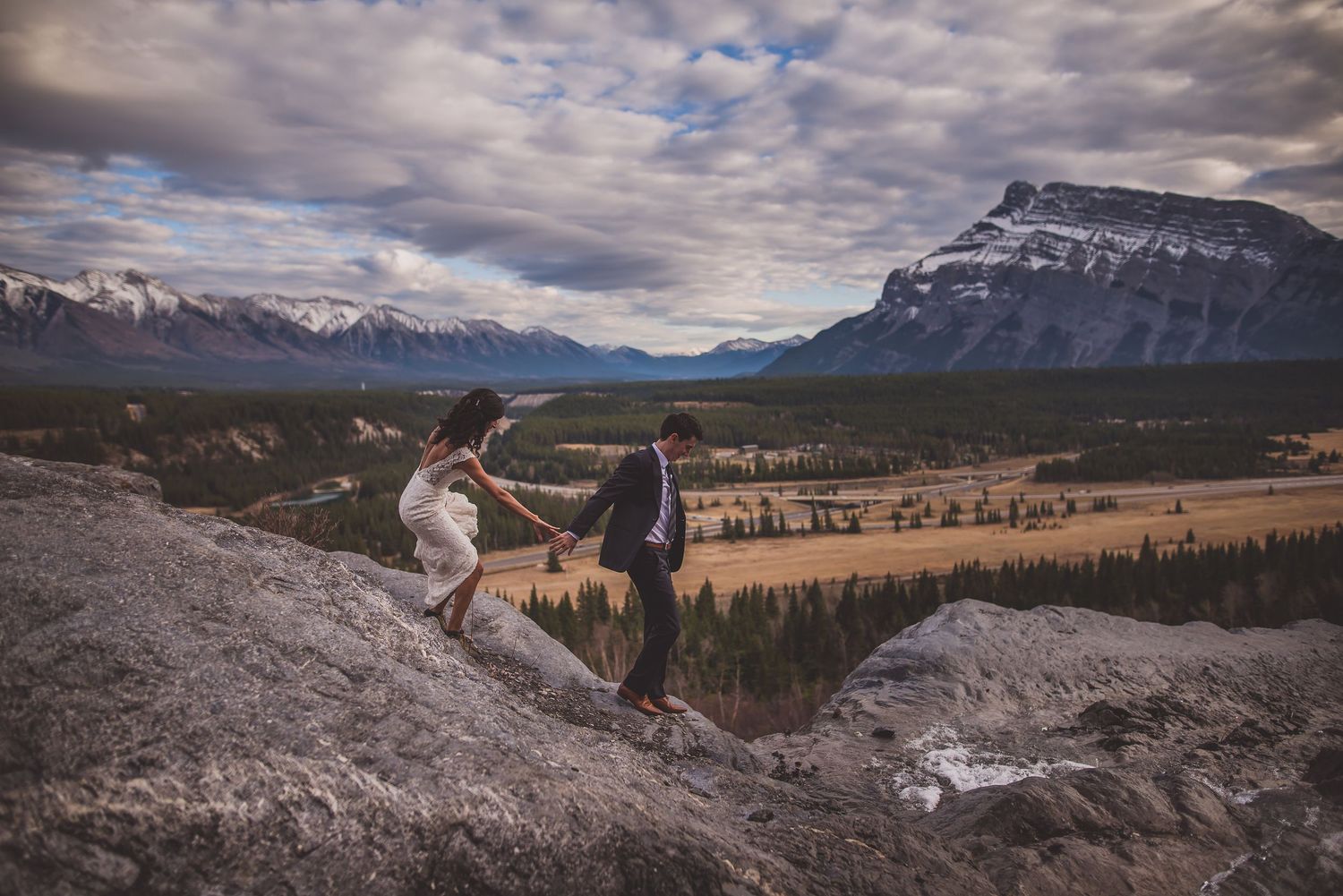 Bride and groom holding hands climbing rocky mountain ridge with stunning Canadian Rockies valley view.