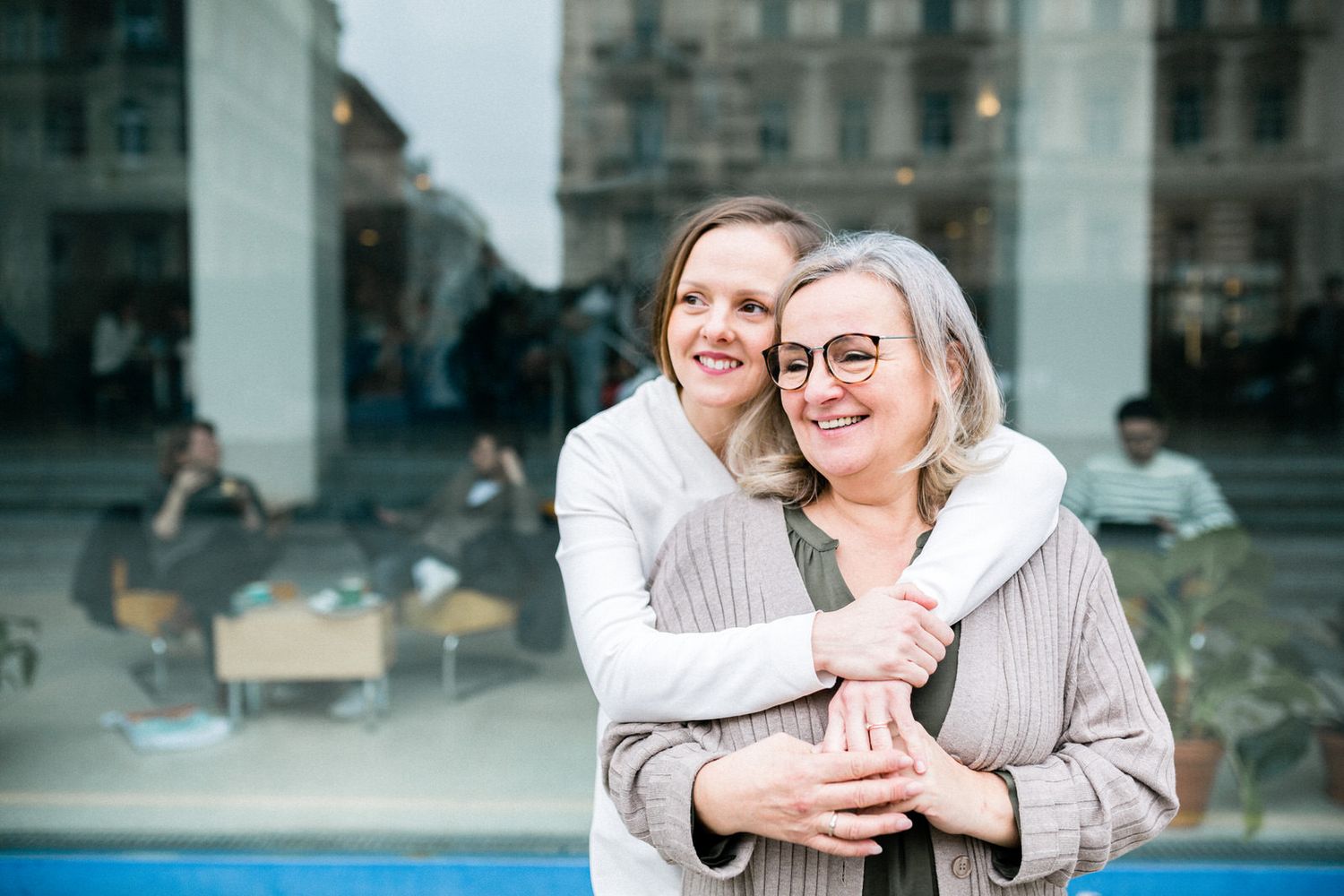Cheerful women share warm embrace outside building with city background.