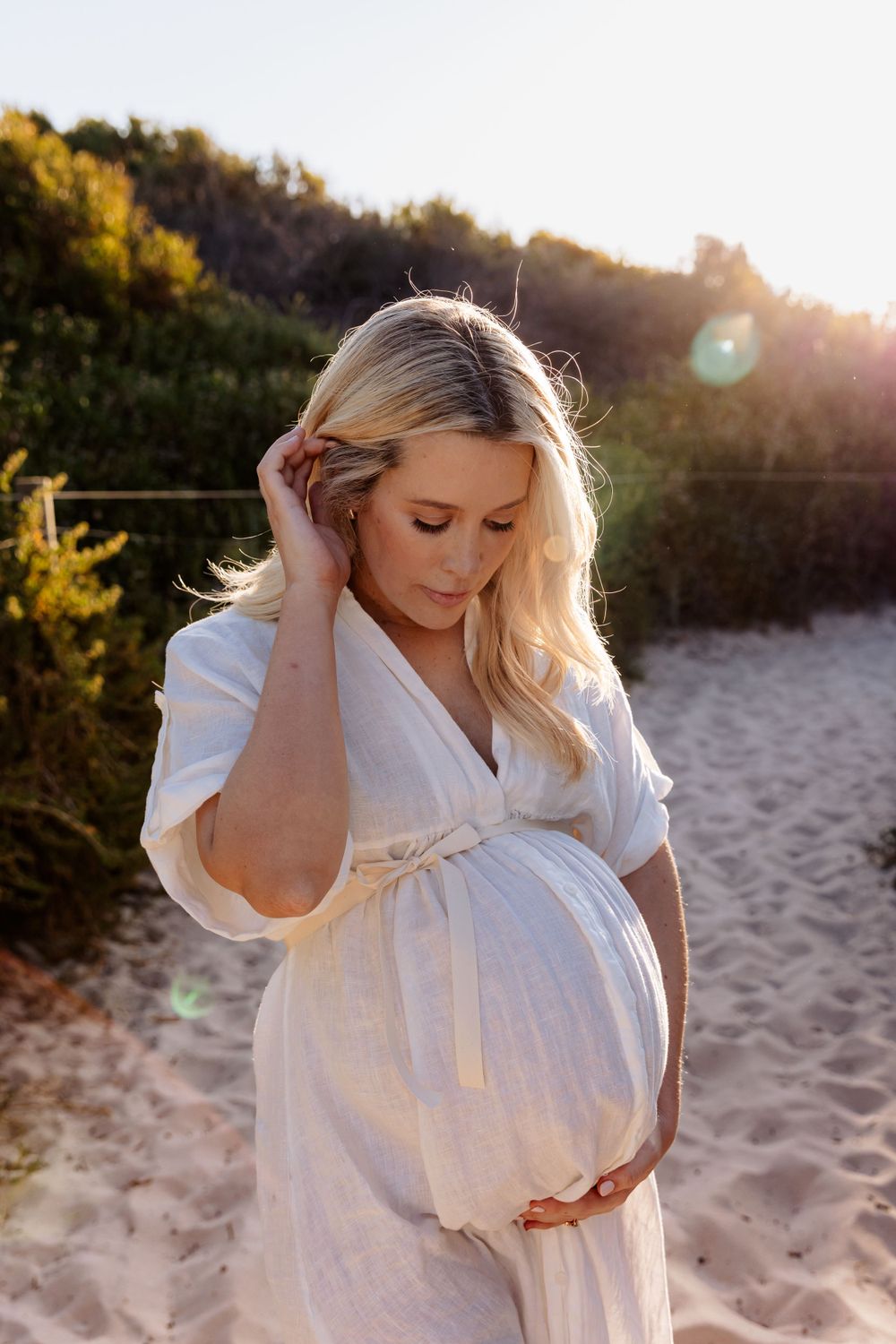 An expectant mother in a white dress poses on a beach at golden hour.