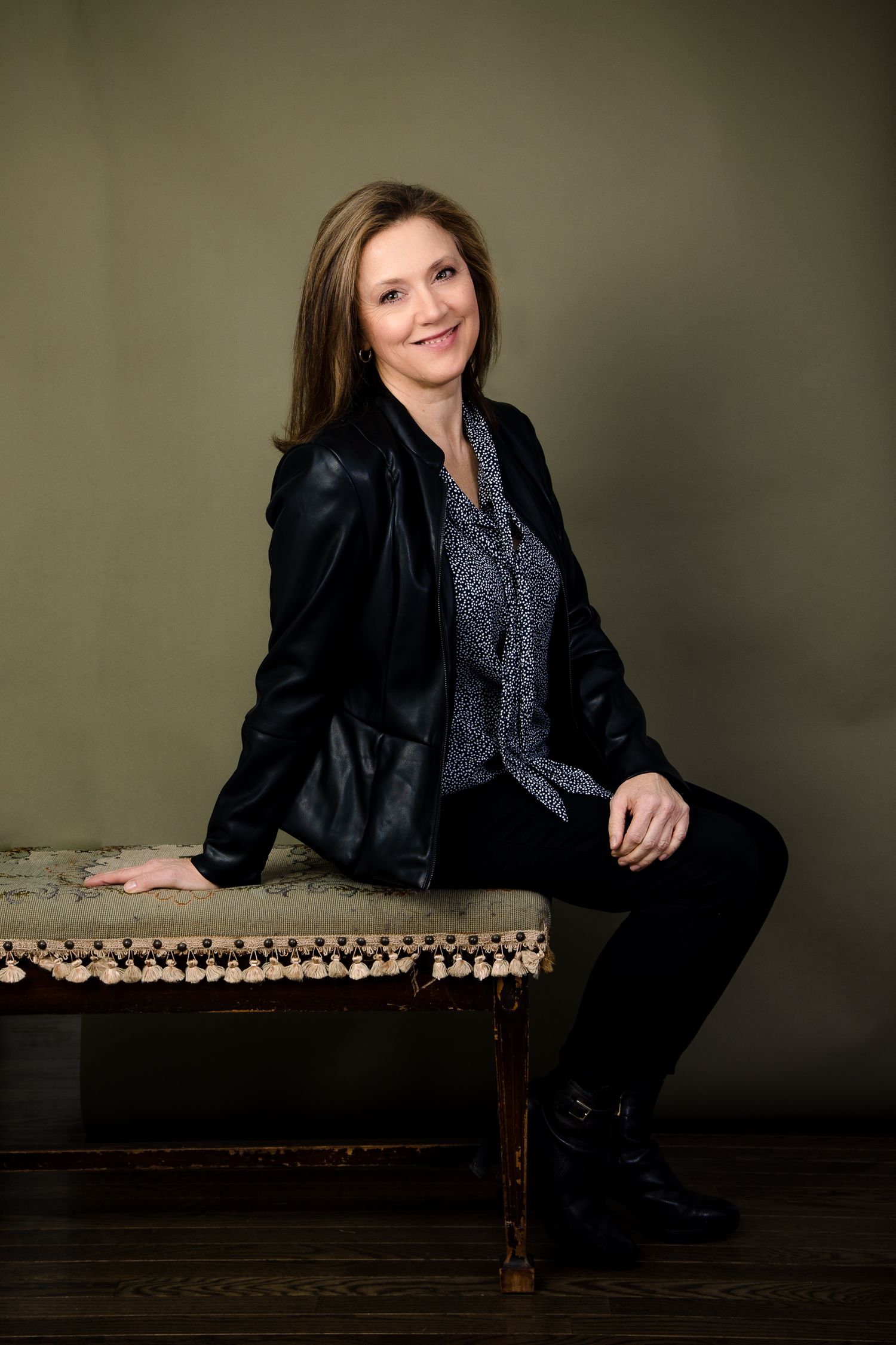 Professional portrait of woman in black blazer sitting casually on antique bench against olive backdrop.