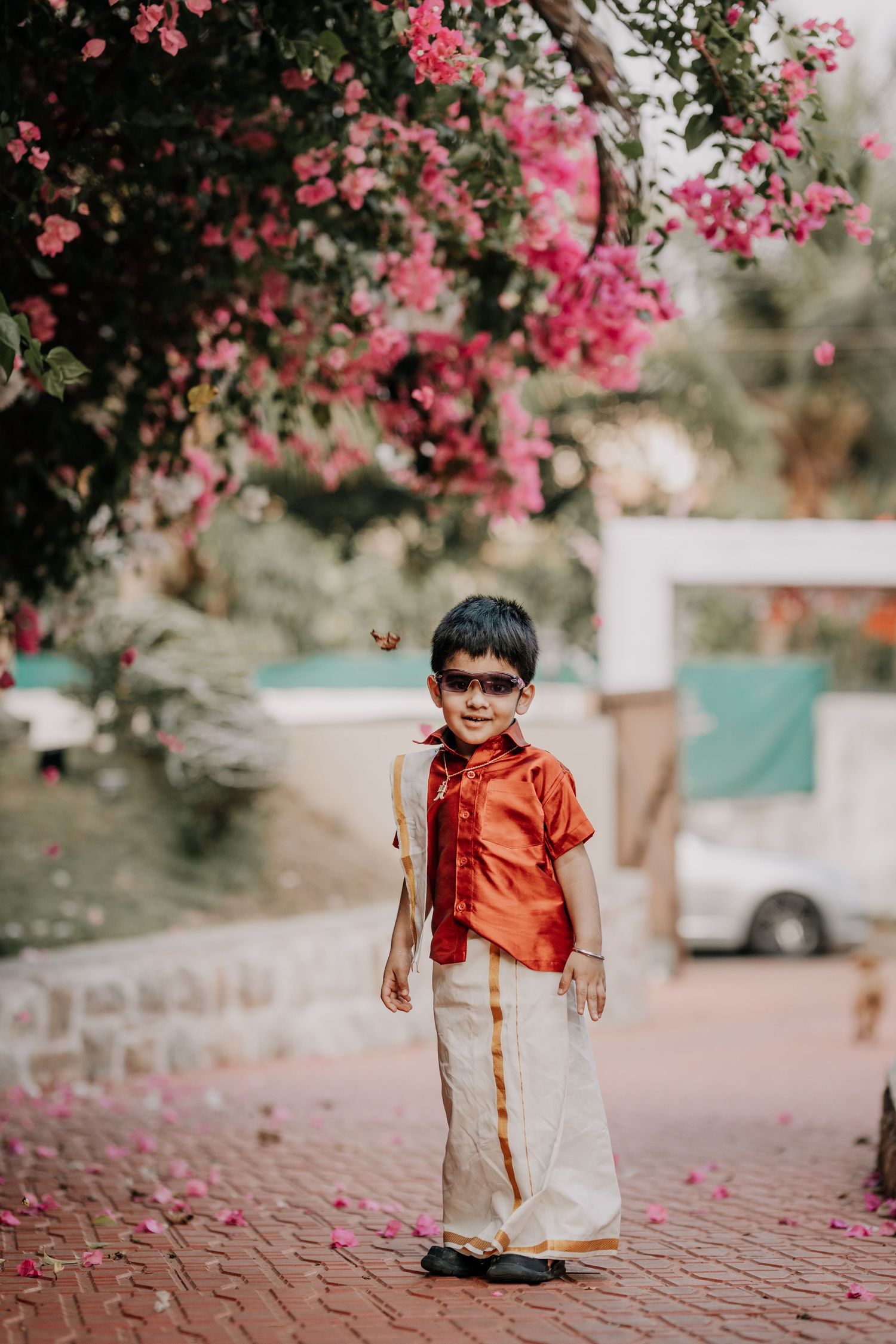 Young child wearing orange and white traditional Indian outfit walking on brick path under pink flowers.