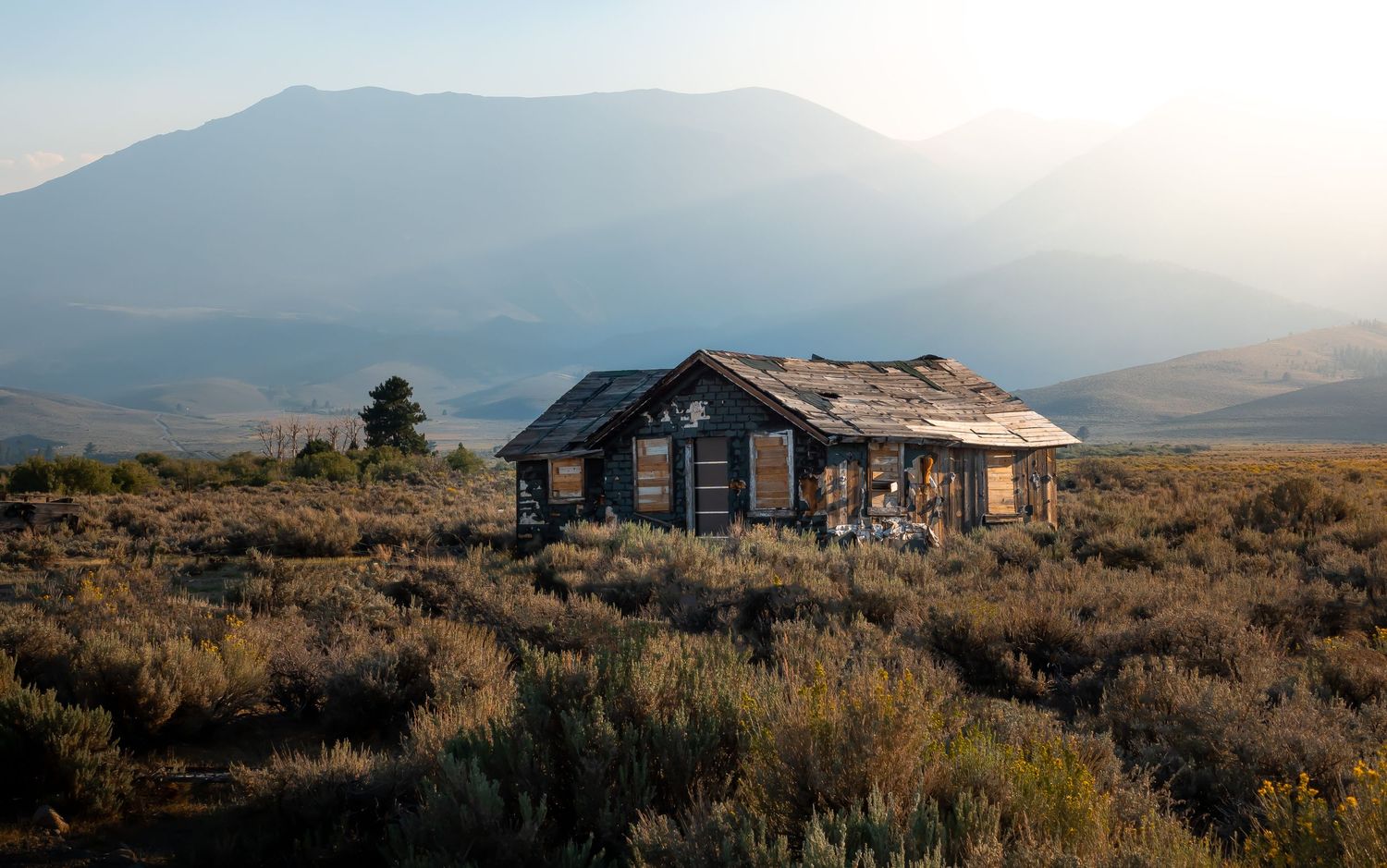 Rustic wooden cabin sits alone on a mountain plain during golden hour with mountains in the background.
