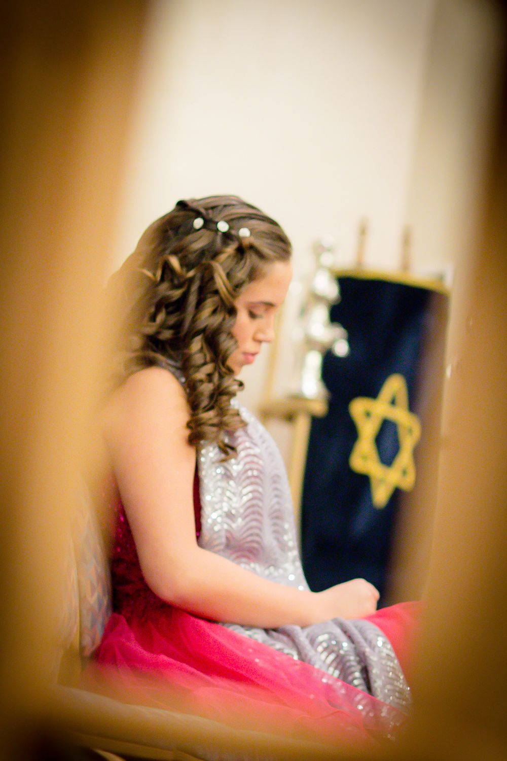 Side profile of someone in formal dress with curled hair sitting near a blue banner with gold Star of David.