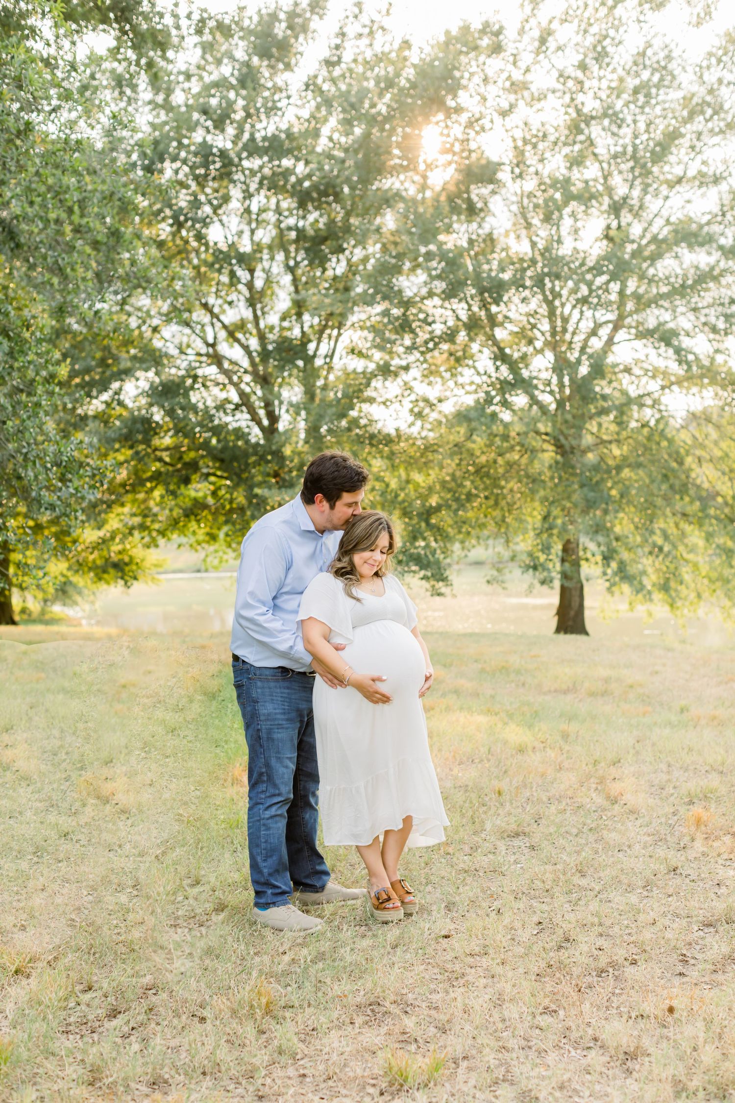A romantic maternity photo of a couple backlit by sunset in a serene outdoor setting.