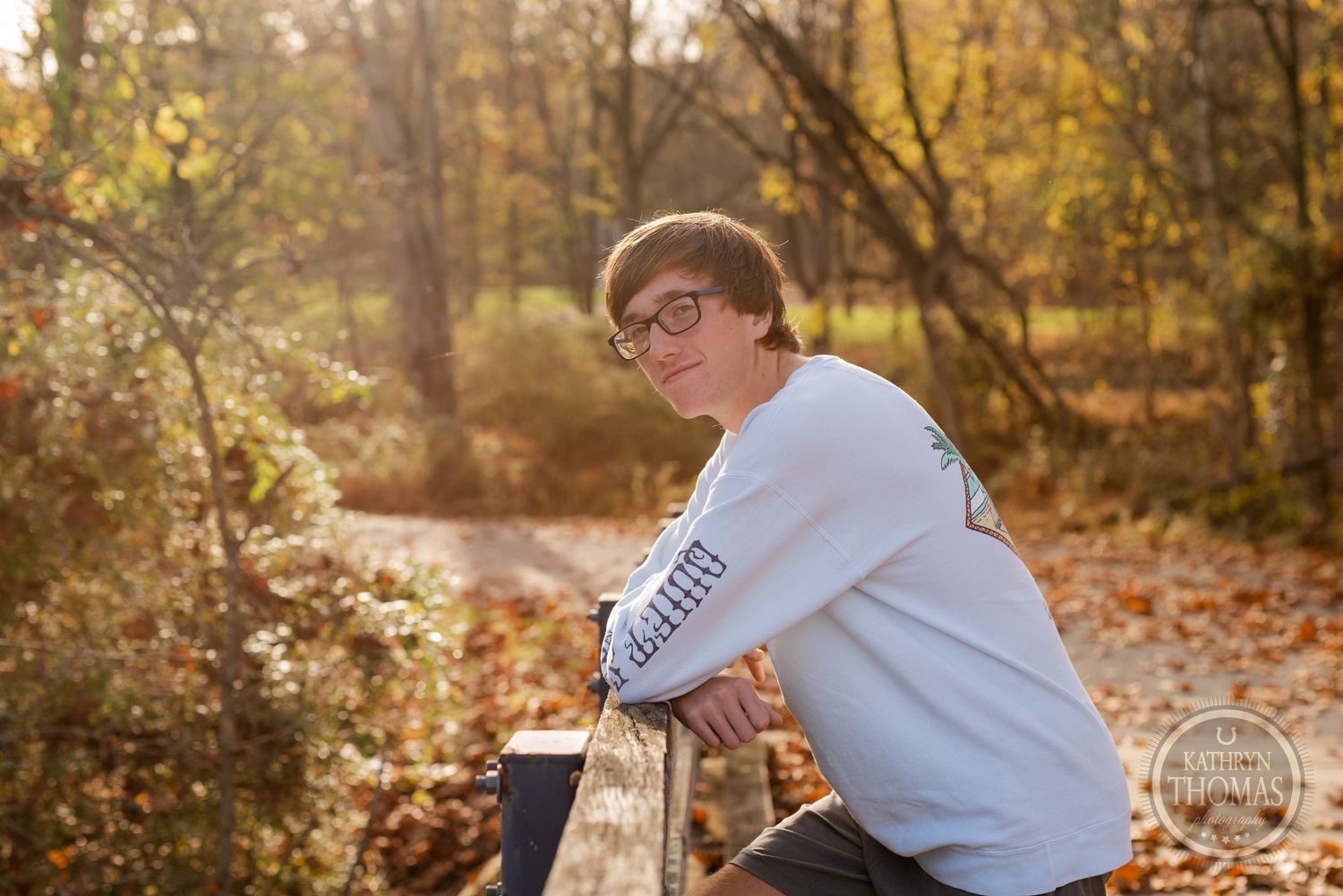 Student athlete in white sweatshirt poses casually on wooden bridge during autumn photoshoot.