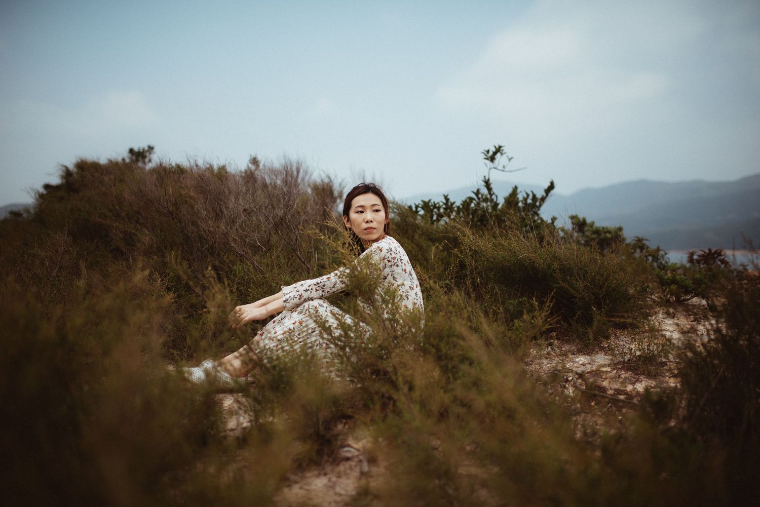a woman sits in the bushes on a hillside looking off into the distance
