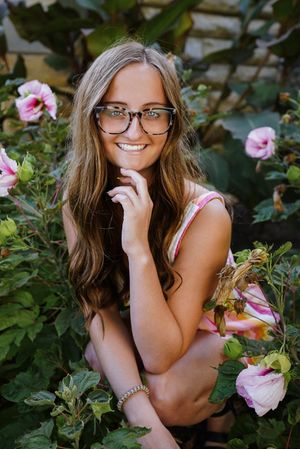 Portrait among pink flowers wearing glasses and pink top in garden setting.