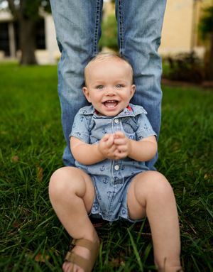 Smiling baby in denim romper sitting between adult legs on grass.