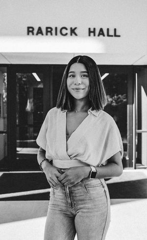 Black and white photo of a person in a light blouse and jeans standing in front of Rarick Hall.