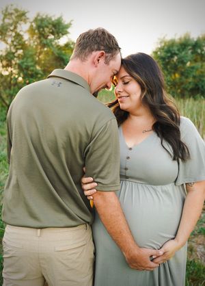An intimate maternity photo shows a couple embracing outdoors, with the woman wearing a sage green dress.