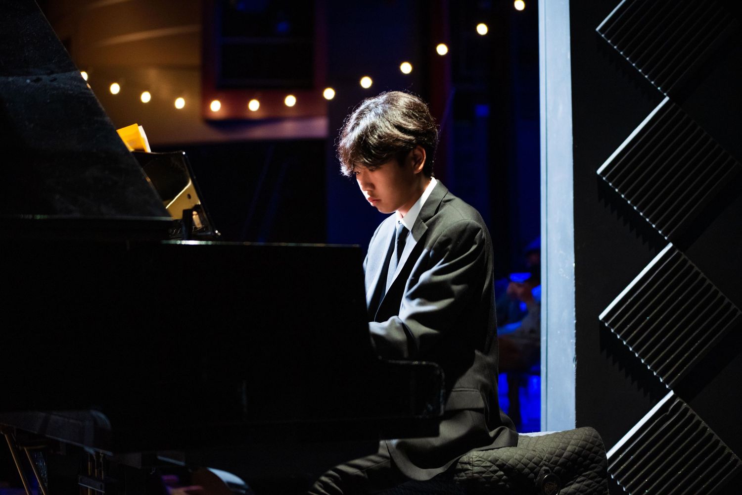 Student playing piano during a concert performance in a theater, with string lights creating bokeh in the background.