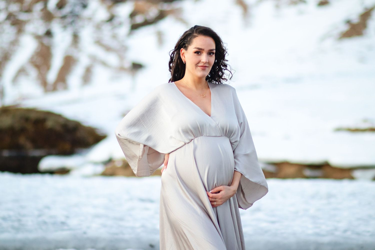 A maternity portrait shows a woman in flowing gray dress against winter coastal landscape.