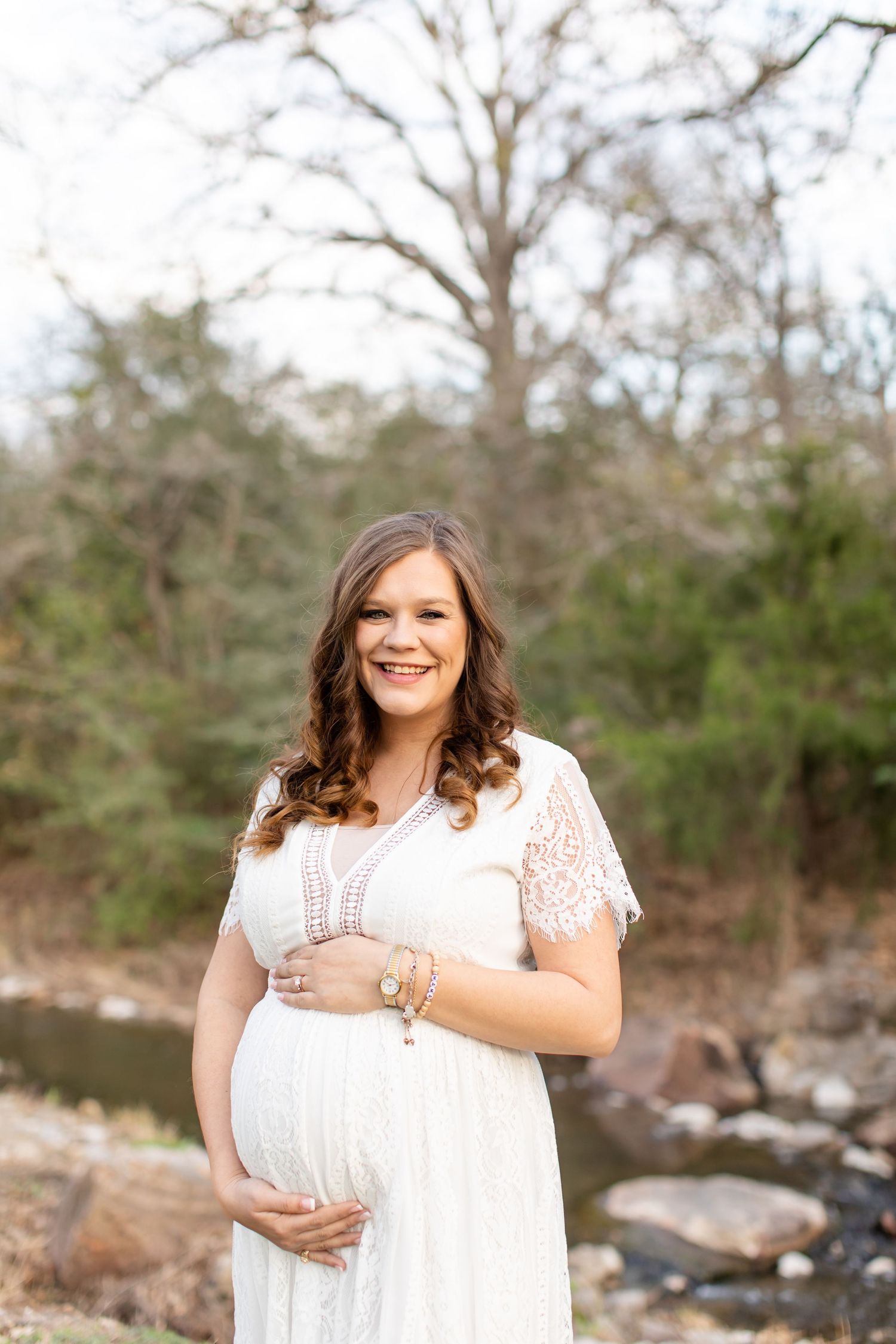 A pregnant woman in a white lace dress smiles while standing near a rocky stream.
