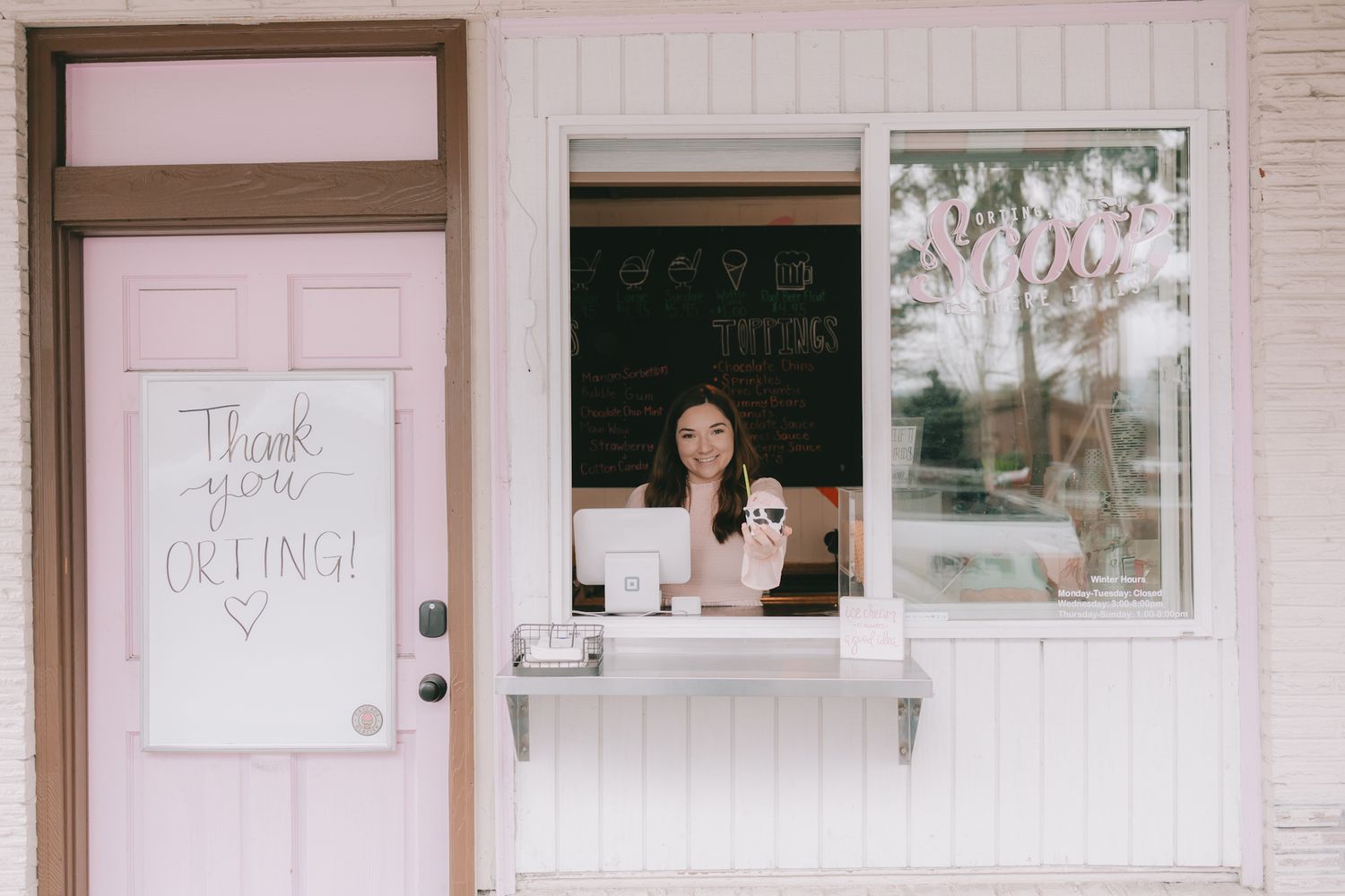 a woman holding an ice cream out of a walk up window the window says Scoop There it is. The pink door whiteboard say thank you orting heart