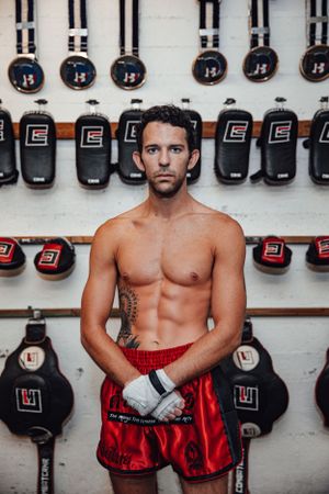 Fighter in red shorts and boxing gloves posing against wall of training equipment.