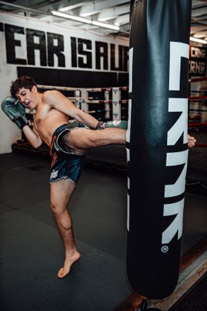 Martial artist practicing high kicks on a heavy bag in a dimly lit gym with multiple training bags visible.