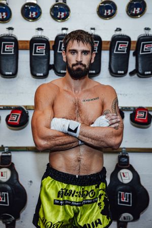 Shirtless fighter with beard poses in yellow shorts against wall of punching bags.