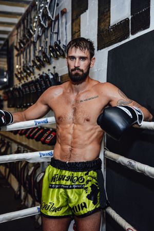 Muscular fighter in neon shorts leans against boxing ring ropes during training.