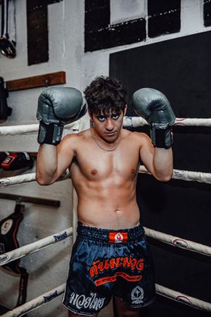 Fighter in blue shorts poses with boxing gloves against boxing ring ropes.