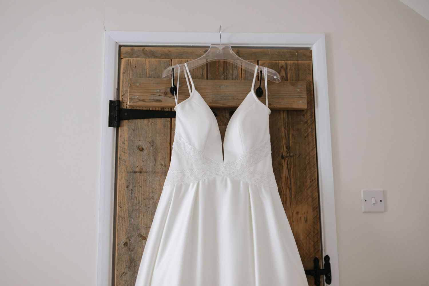 An elegant white wedding dress with spaghetti straps hanging on a wooden door.