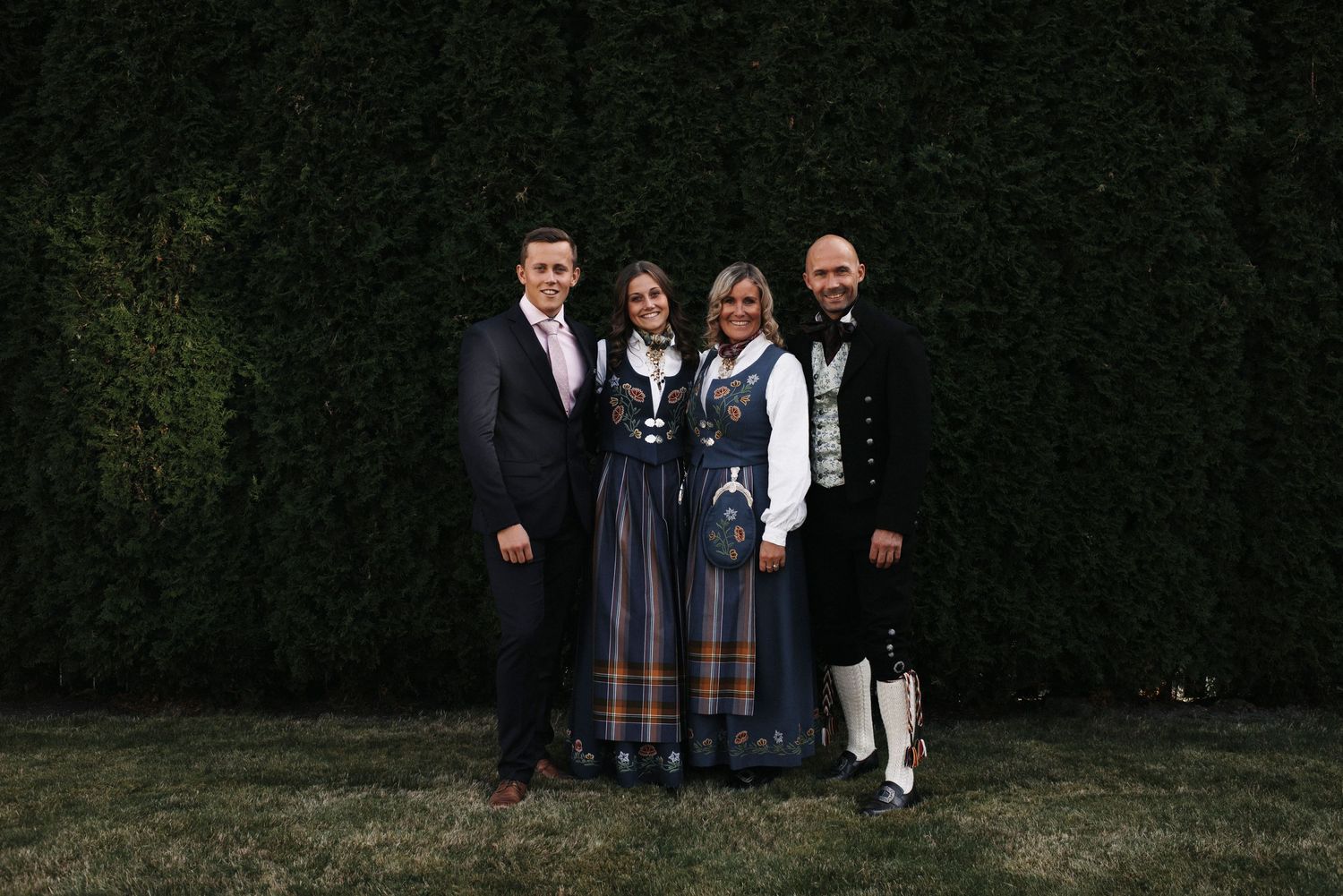Family group photo in traditional Norwegian formal wear and bunads against a green hedge background.