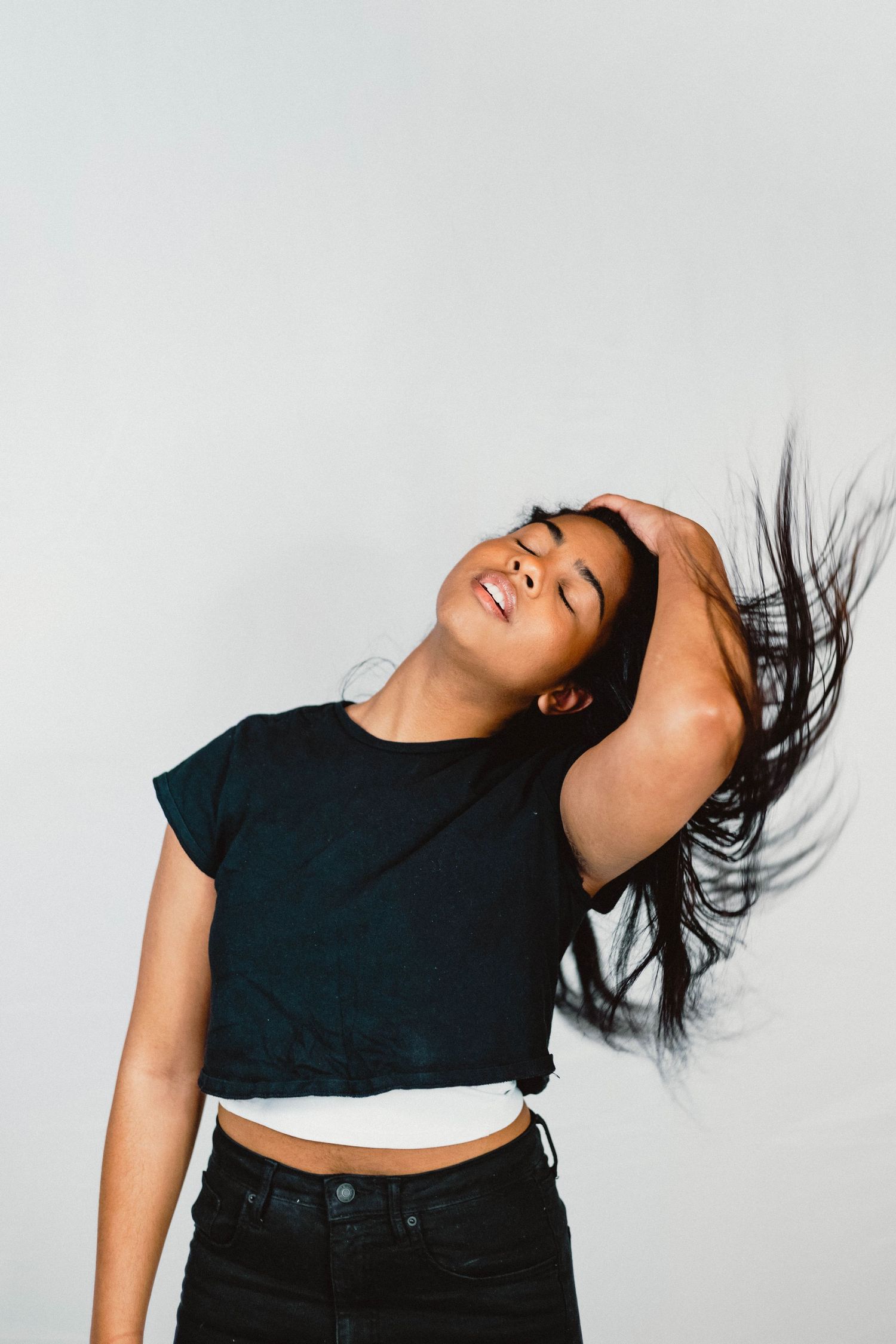 A person in a black crop top and dark pants flips their hair while dancing against a white background.