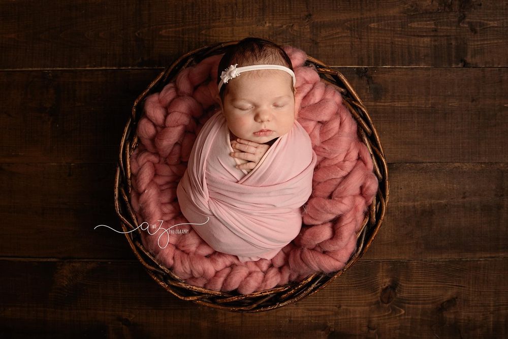 Sleeping newborn wrapped in pink fabric nestled in round basket with rosy padding.