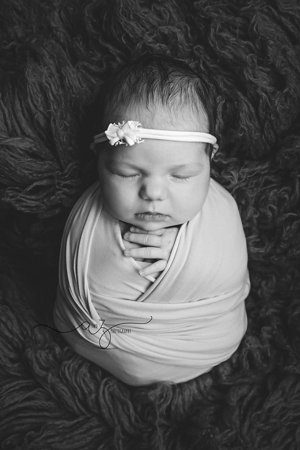 Black and white portrait of newborn wrapped in fabric with delicate headband on textured background.