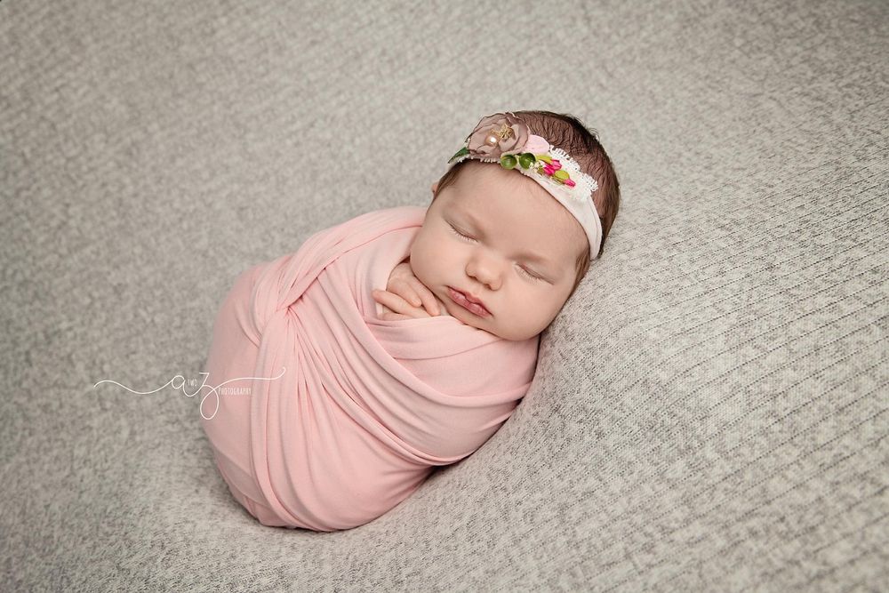 Newborn baby sleeping peacefully in pink wrap with floral headband on grey background.