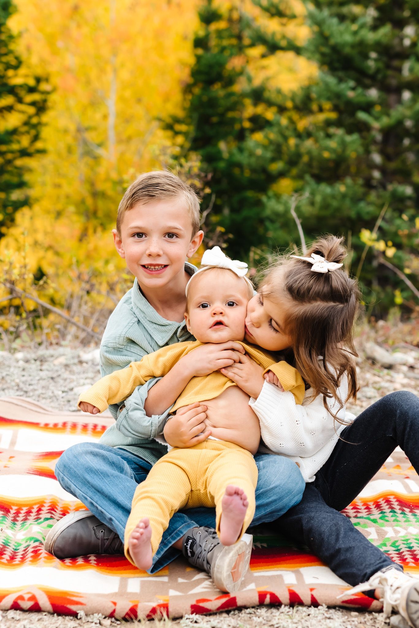 Children playing and hugging on a red and white blanket with vibrant yellow fall foliage in the background.