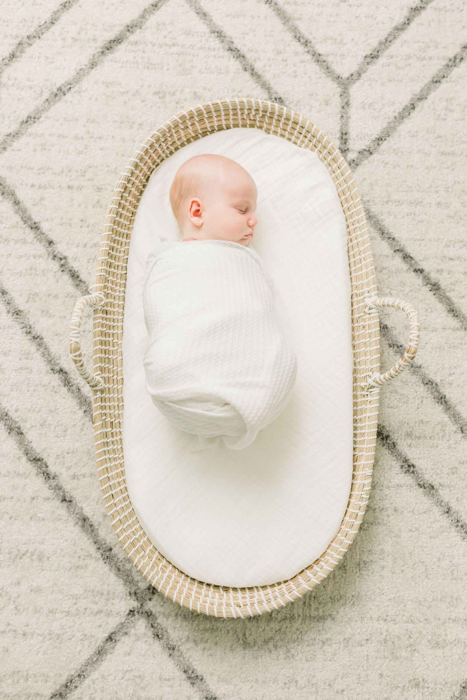 Infant wrapped in white blanket resting peacefully in oval woven moses basket.