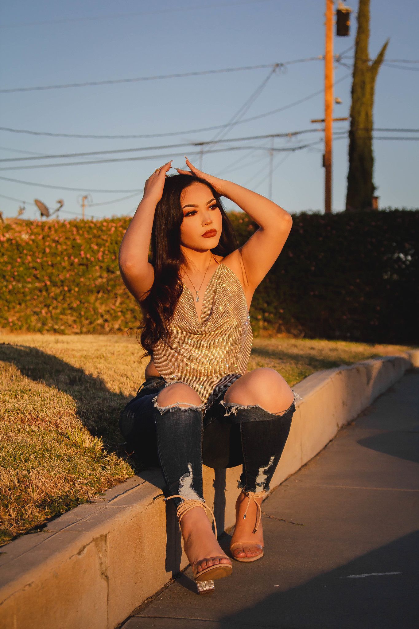 Woman in sparkly top and ripped jeans poses at sunset on concrete ledge.