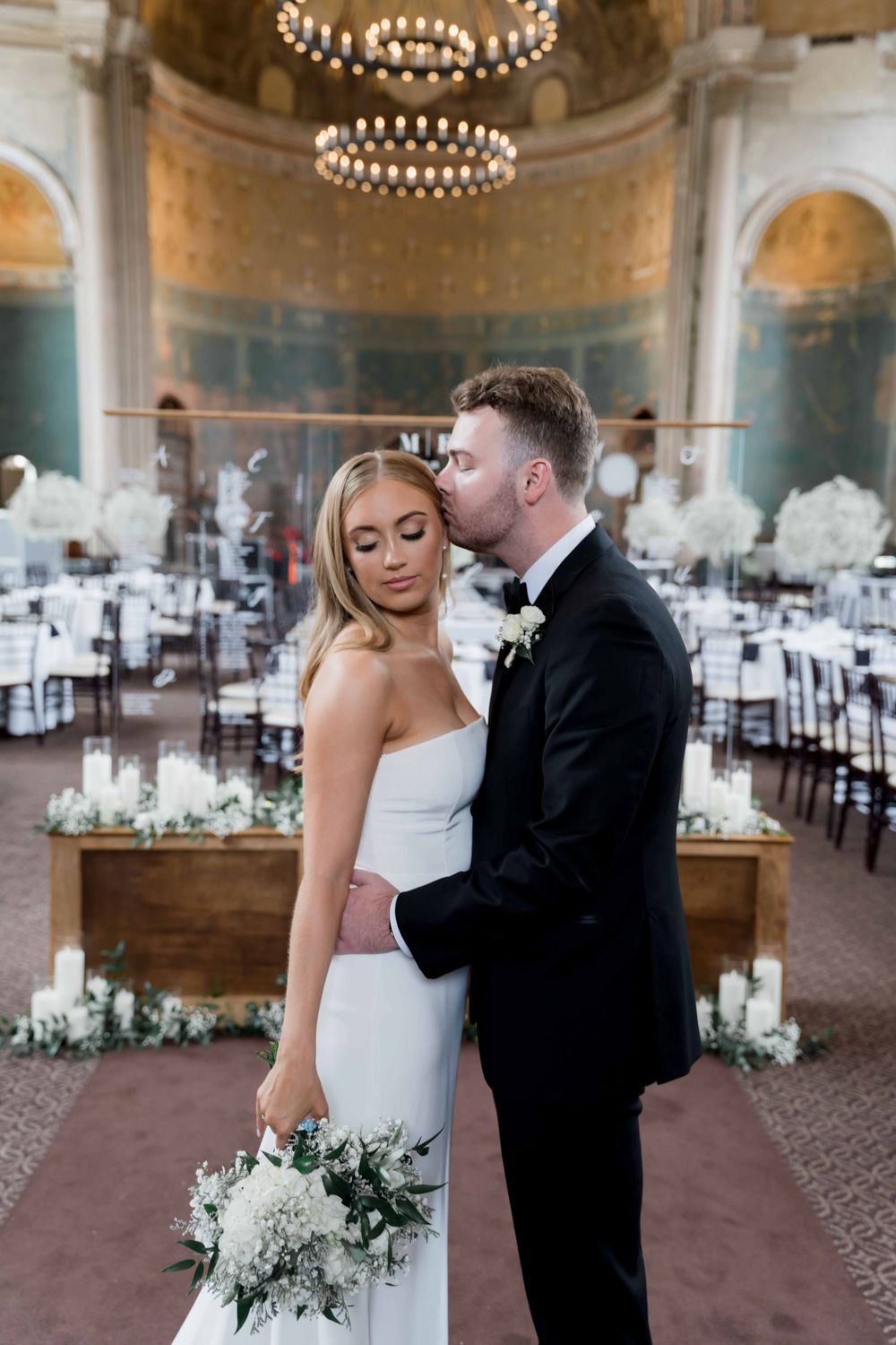 Romantic moment between newlyweds in an elegant ballroom with chandeliers and floral decorations.