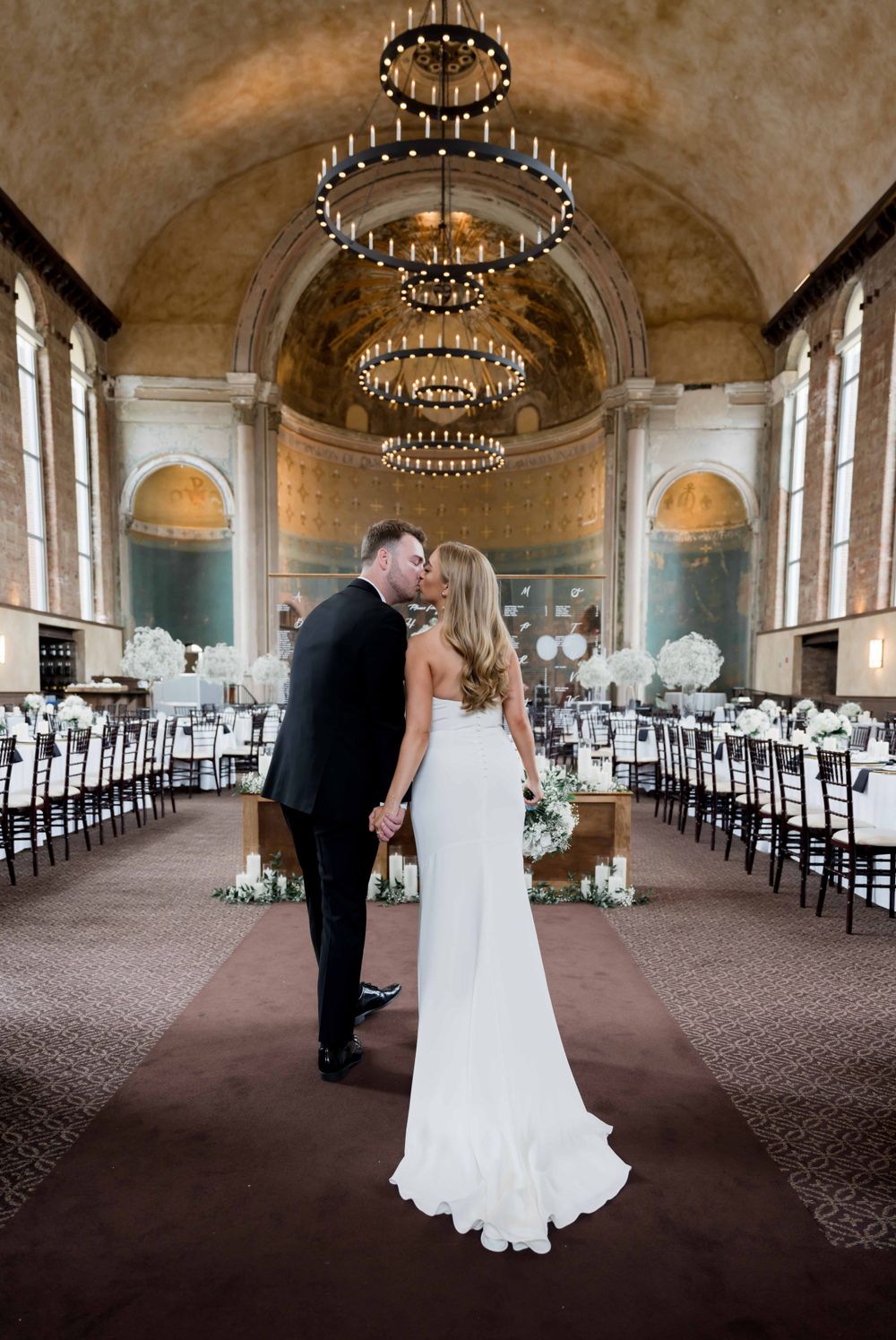 Newlyweds share an intimate moment in a grand ballroom with stunning chandeliers and table settings.