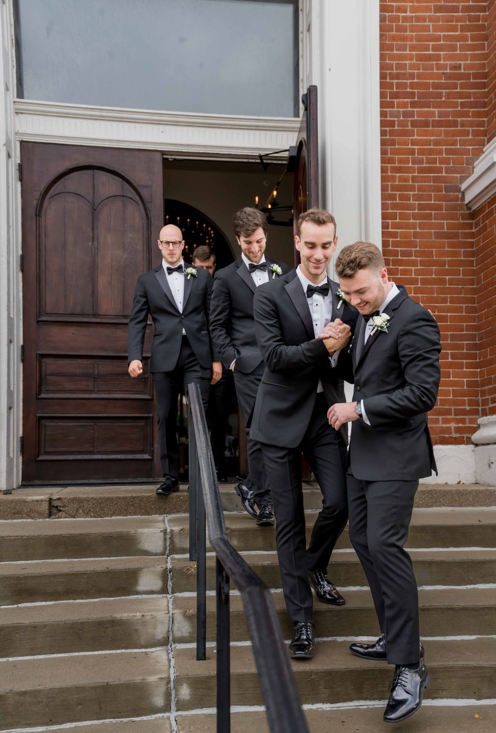 Groomsmen in black tuxedos descending church steps with brick facade in elegant wedding party entrance.