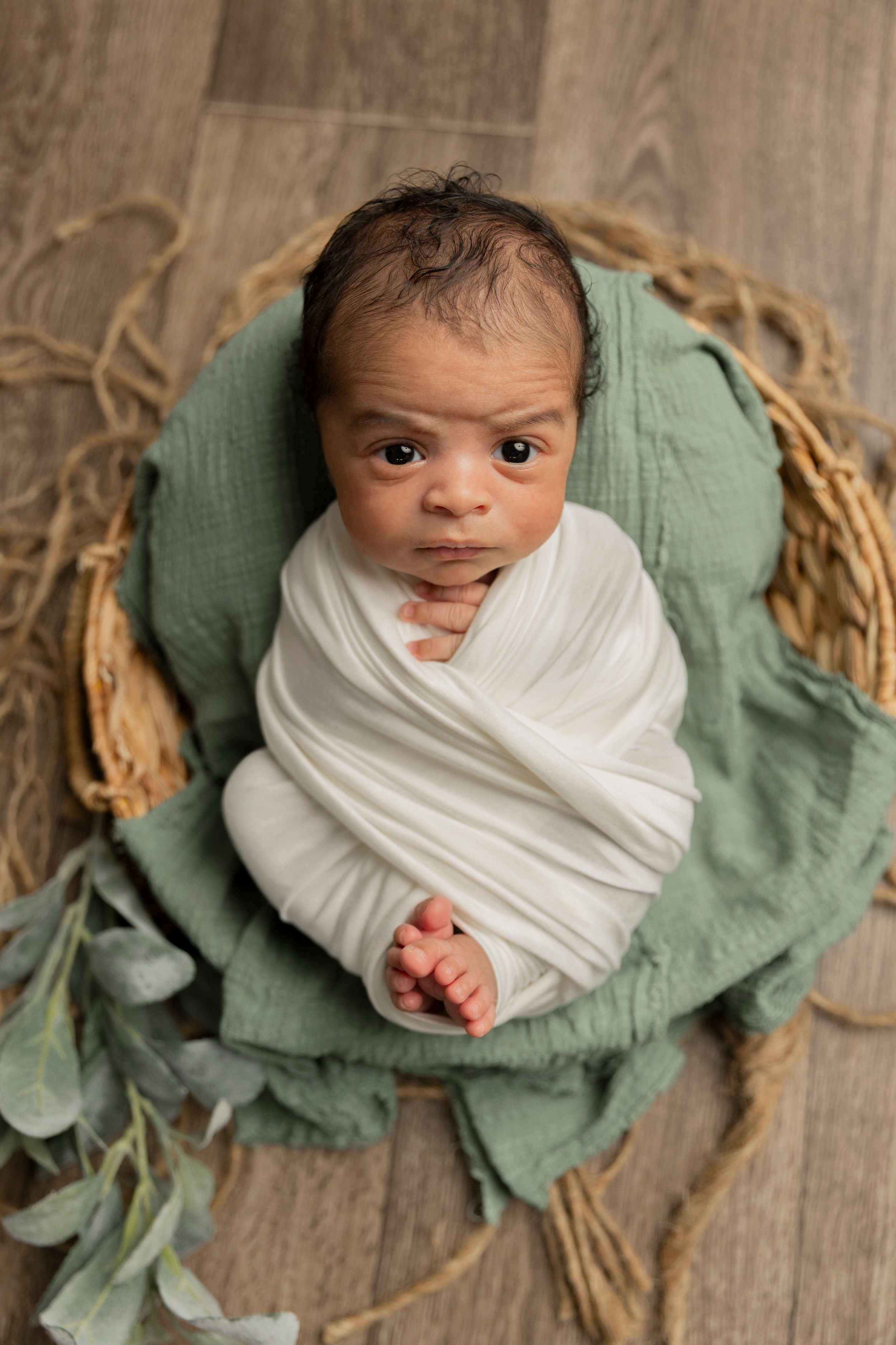 newborn baby wrapped in white with a sage green background looks at the camera while his toes and fingers and showing