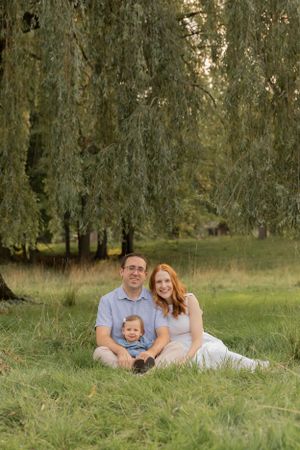 family of three sitting in a field in front of a willow tree for a photo
