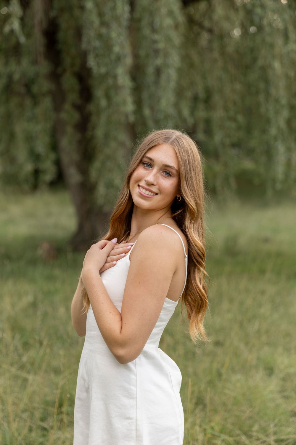 teenage girl wearing a white dress smiles for a photo under a willow tree in Oakland county michigan