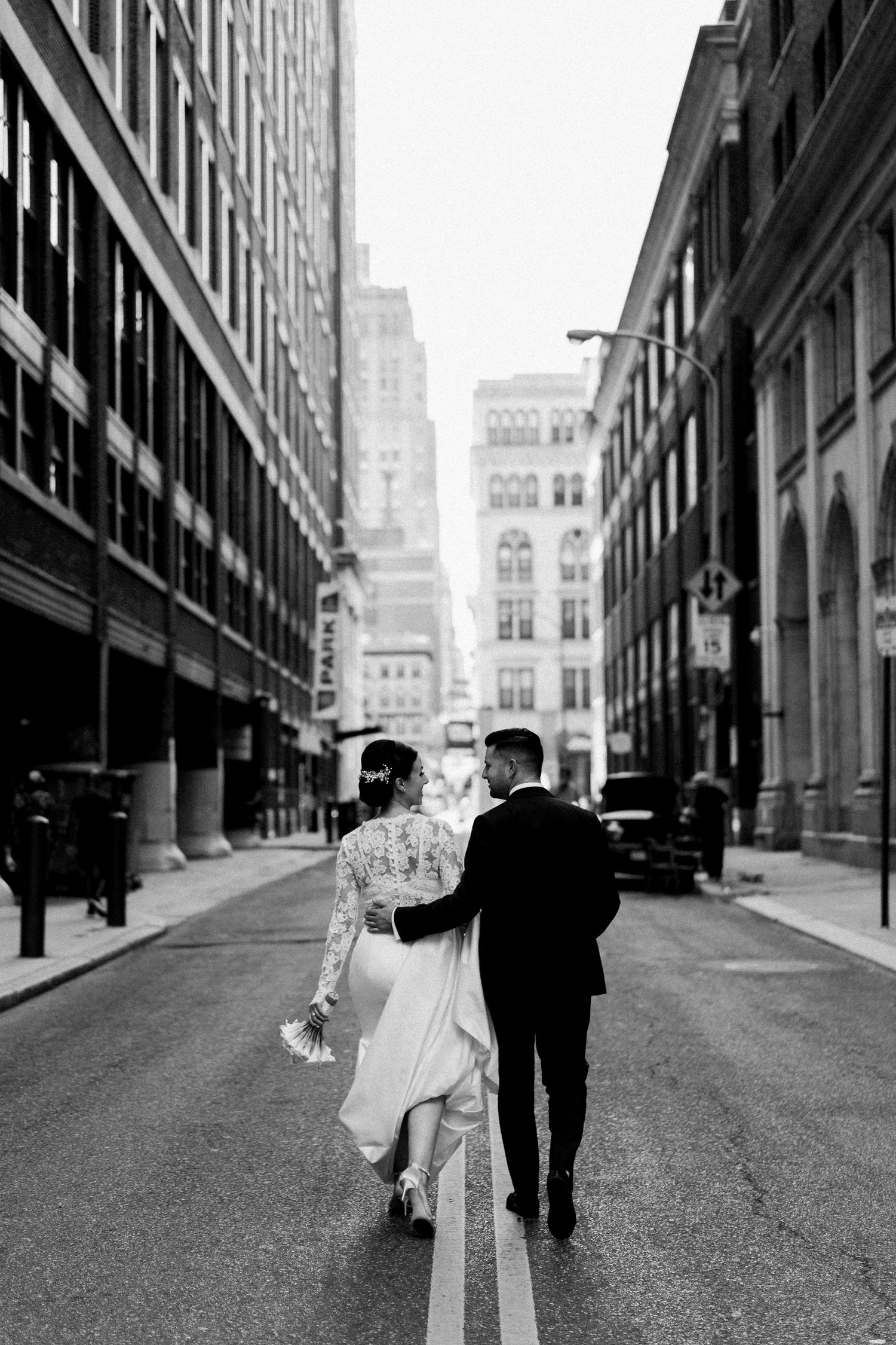Black and white photo of newlyweds walking down a city street between tall buildings.