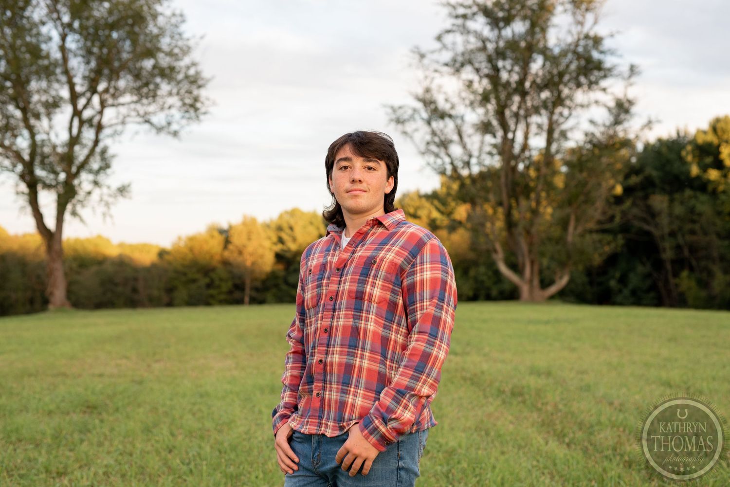 Person in a plaid shirt stands in grassy field at sunset with trees in background.