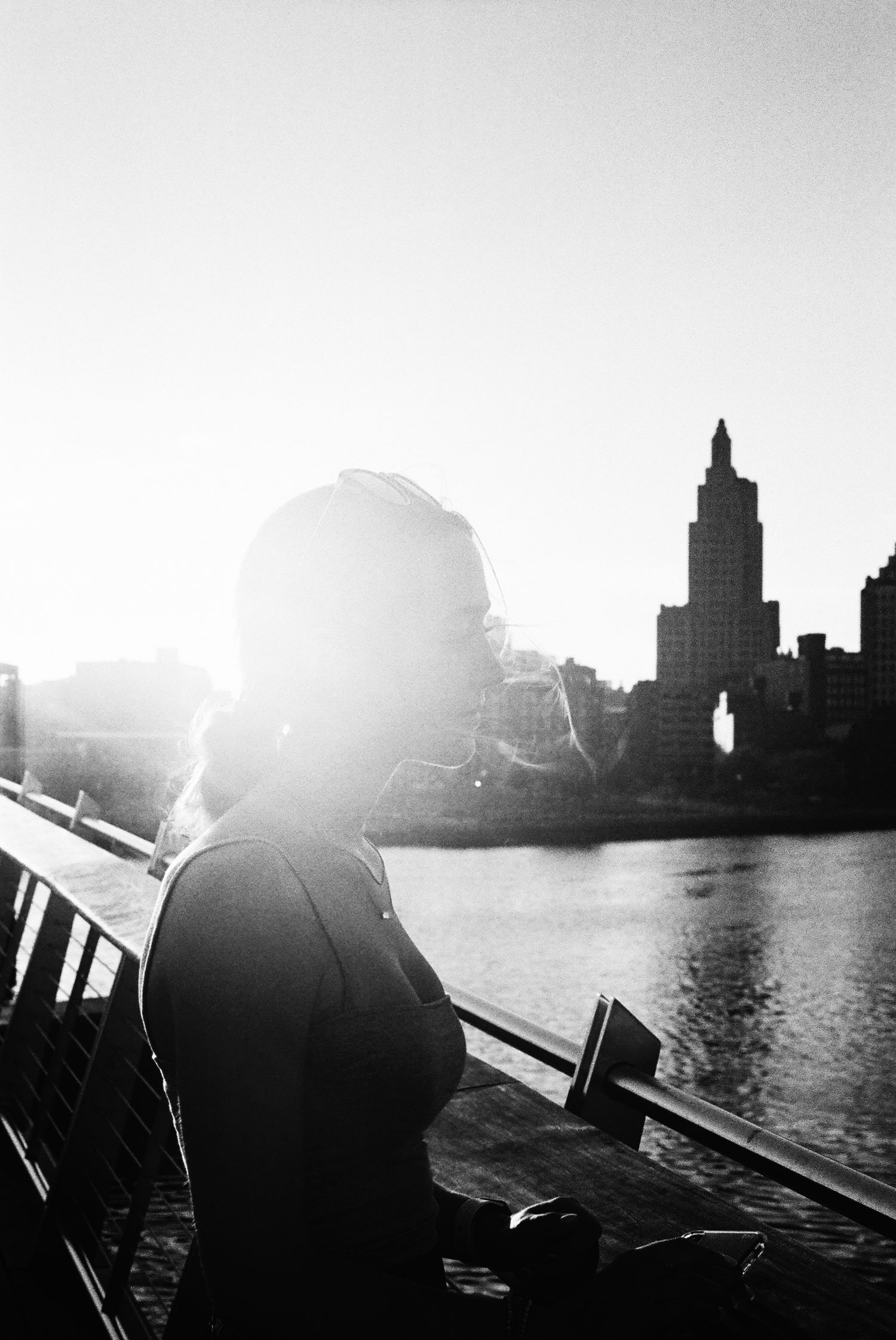 Silhouetted figure watches sunset over city skyline with tall buildings reflected in water.
