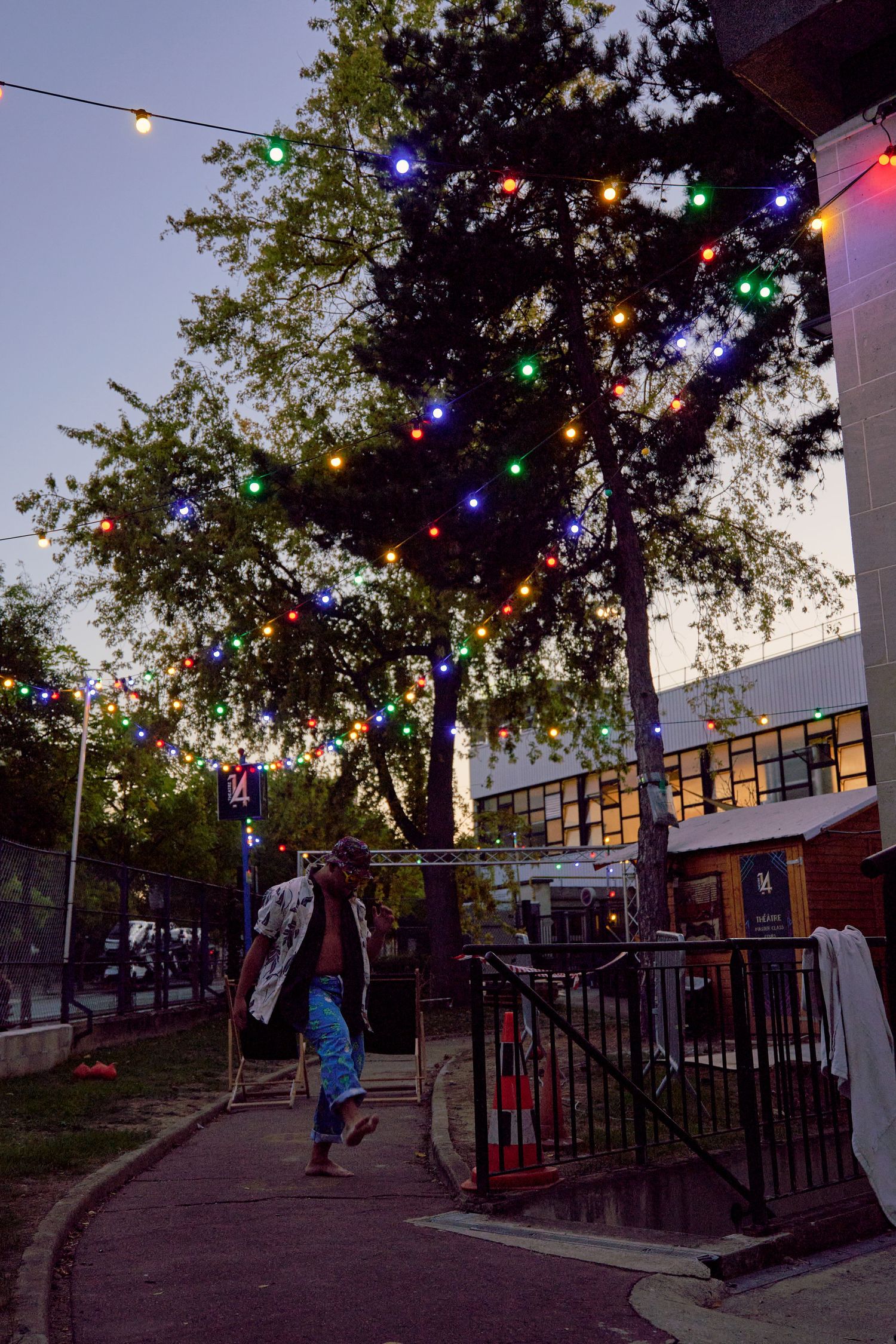 Colorful string lights illuminate trees at dusk in an urban outdoor space.