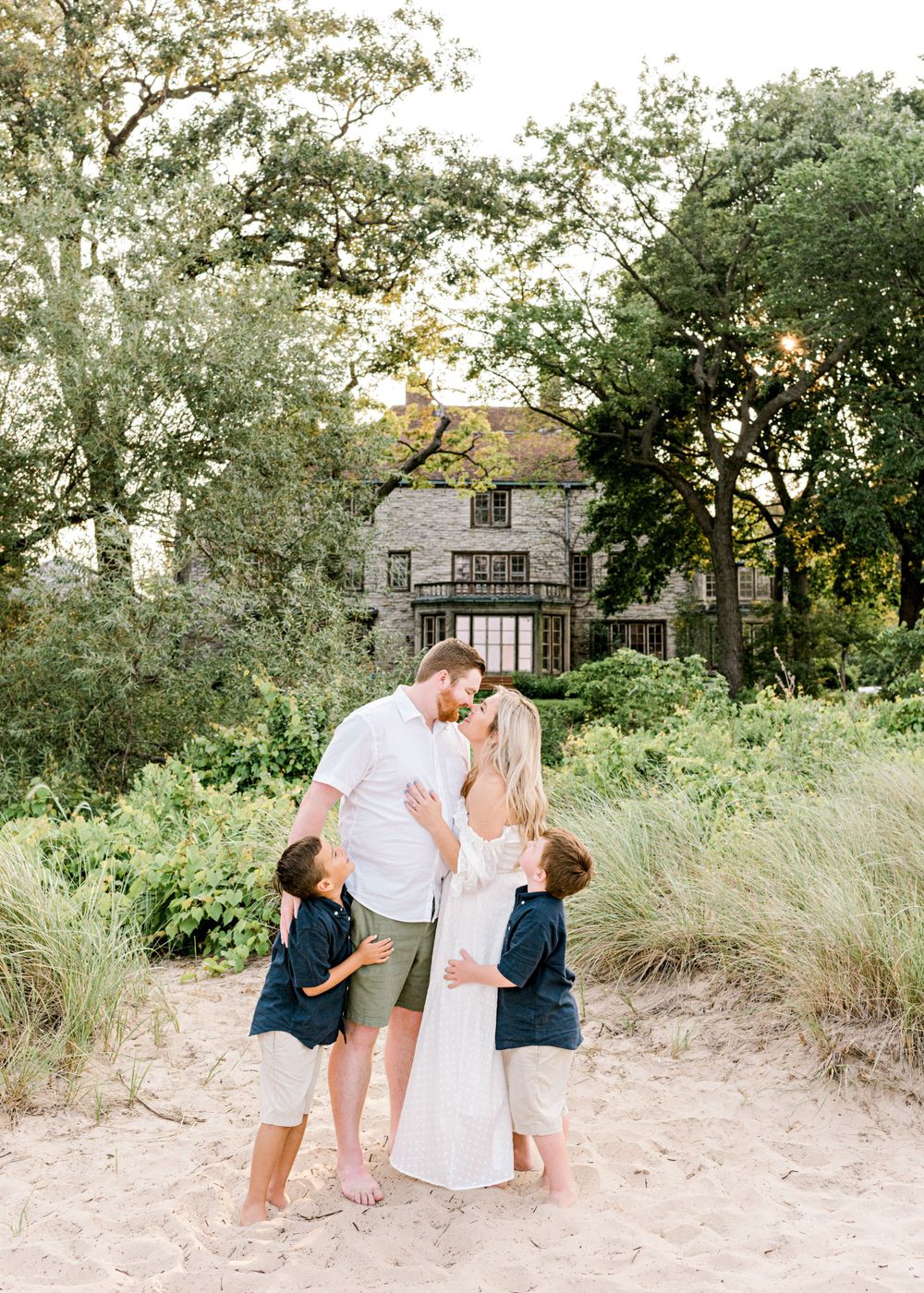A loving family embraces on a sandy beach path with a beautiful house in the background.
