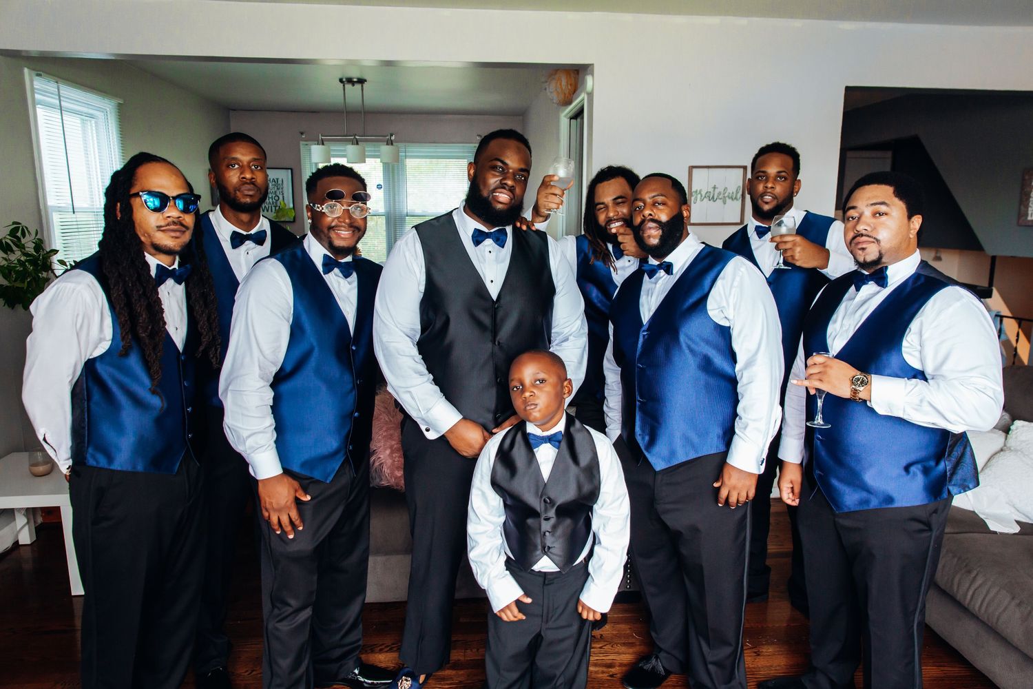 Groomsmen in navy blue vests and bow ties pose with groom in gray vest for wedding party photo indoors.
