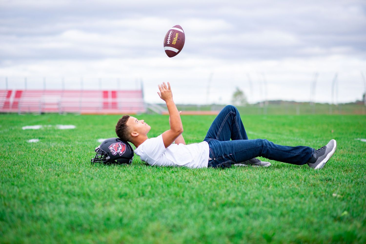 Highschool Senior laying in the grass on his football field while tossing his football in the air and catching it.